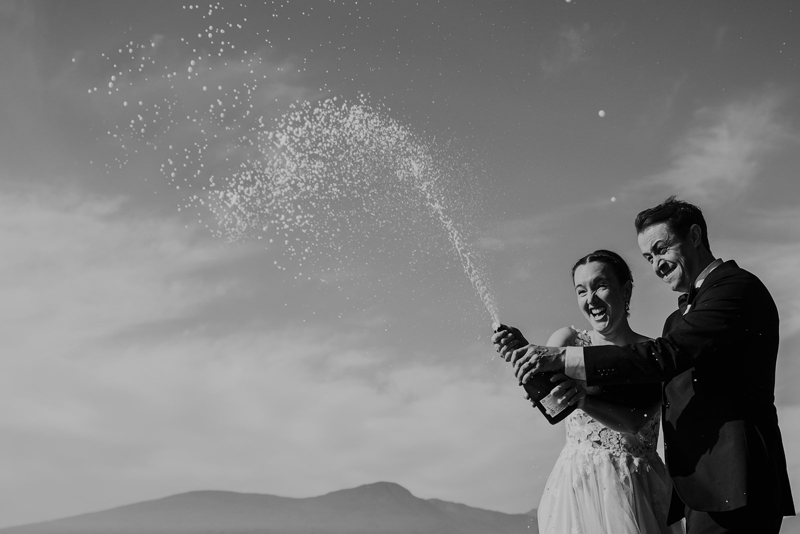 A black-and-white photo of a joyful bride and groom opening a champagne bottle outdoors, photograph taken during their elopement on Bowen Island.